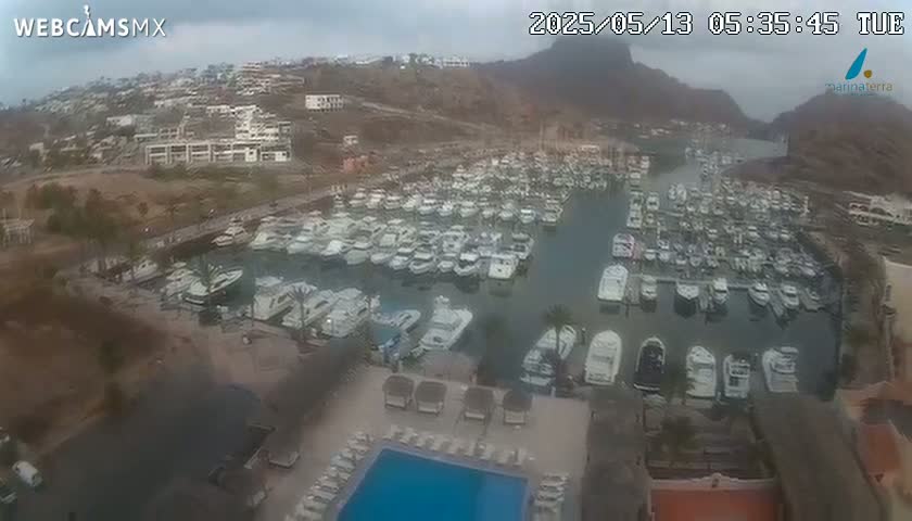 An overcast day reveals a marina filled with numerous boats, nestled beside a resort with a swimming pool and buildings overlooking a hillside.