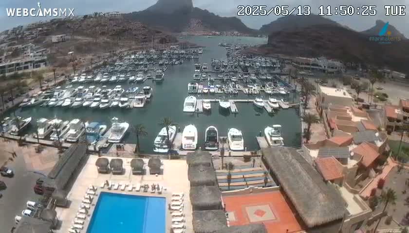 An aerial view of a marina filled with boats on a partly cloudy day, overlooking a resort with a swimming pool and surrounding buildings nestled against a mountainous backdrop.