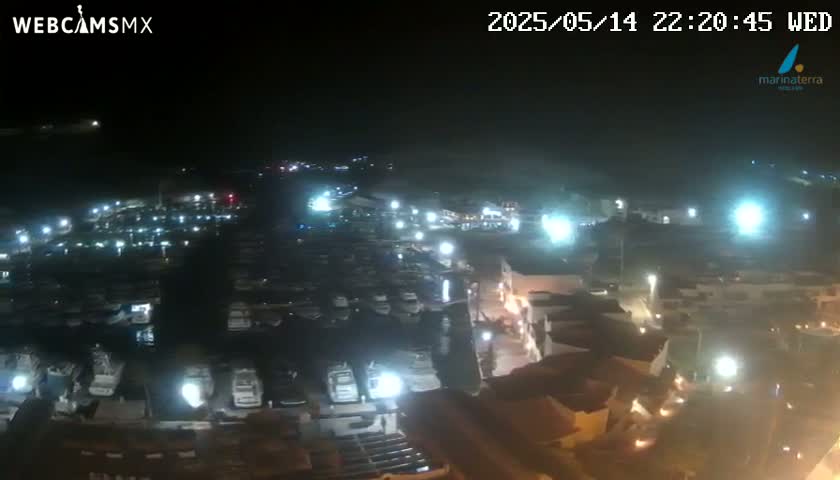 A nighttime aerial view of a marina filled with boats, illuminated by artificial lights, with a town visible in the foreground.
