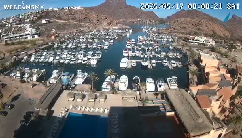 An aerial view shows a marina filled with numerous boats, situated near buildings and a swimming pool, under a clear sunny sky.