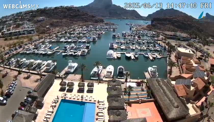 An aerial view shows a marina filled with boats, a resort with a pool, and surrounding desert landscape under a sunny sky.