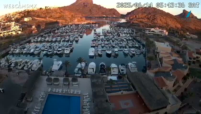 An aerial view of a marina filled with numerous boats, situated near buildings and a swimming pool, under a clear morning sky with a large mountain in the background.