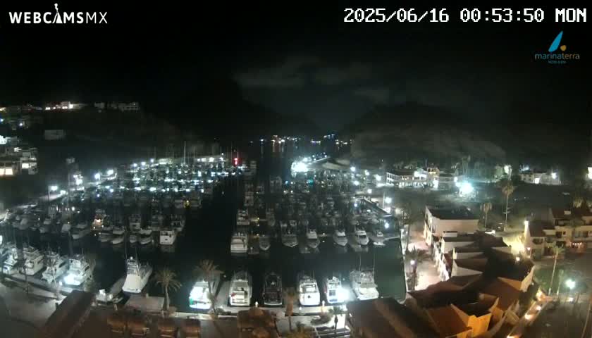 A nighttime aerial view of a marina filled with boats, nestled between hills under a dark, cloudy sky.