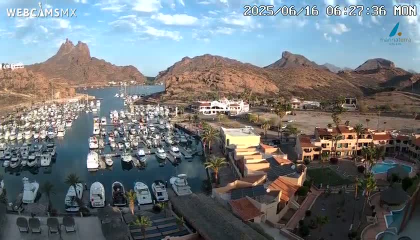 A marina filled with numerous boats is nestled between arid, rocky hills under a mostly clear, sunny sky.