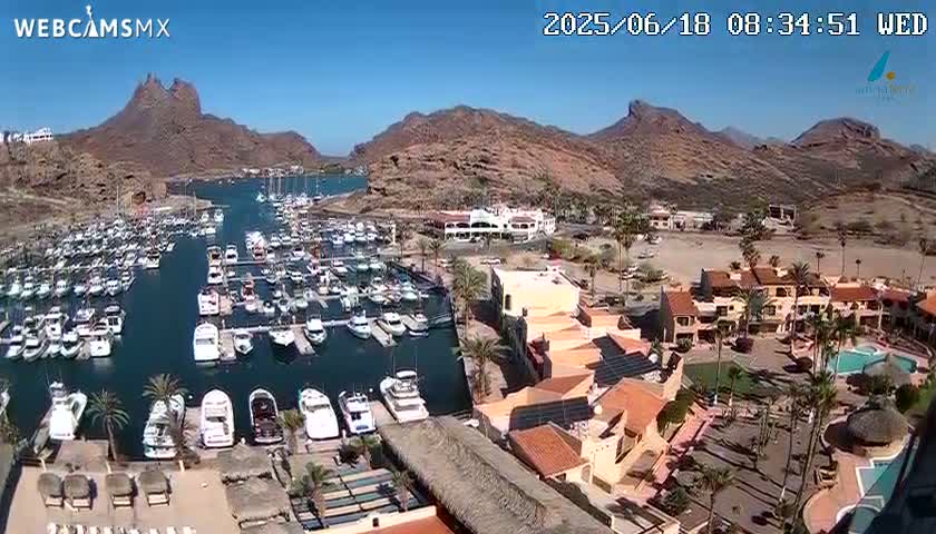 A marina filled with boats is situated next to a resort complex, with mountains in the background under a clear, sunny sky.