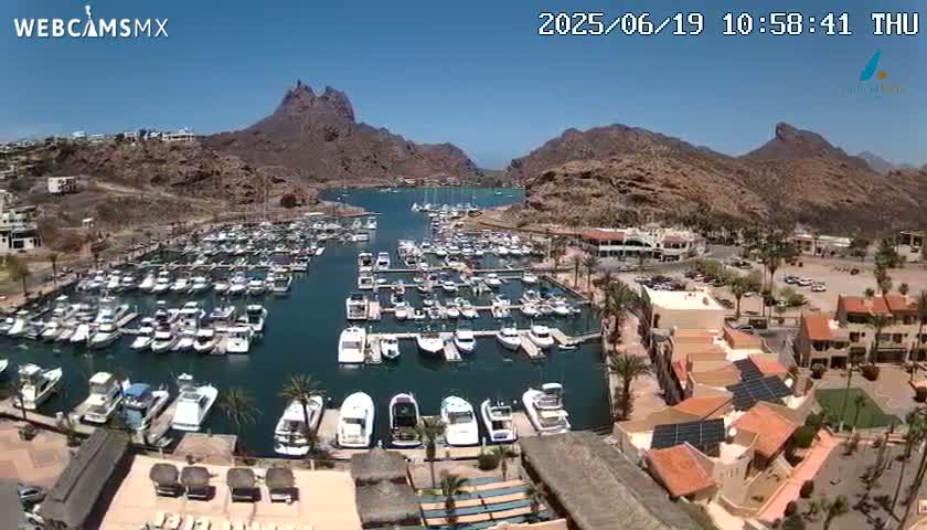 A marina filled with numerous boats is nestled between brown, rocky hills under a clear, sunny sky.