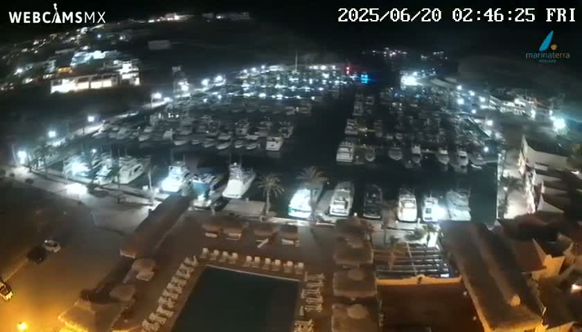 A nighttime aerial view of a marina filled with boats, adjacent to a building with a pool and outdoor seating area under clear skies.
