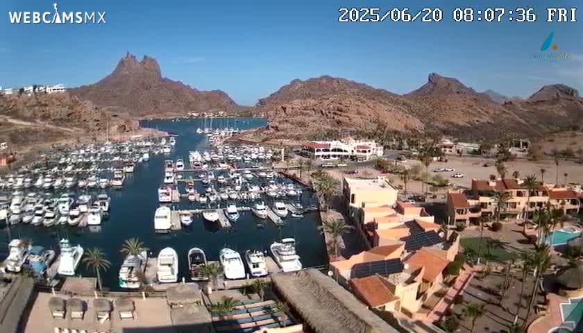 A marina filled with numerous boats is nestled between arid, mountainous terrain under a clear, sunny sky.