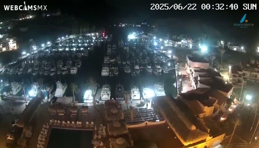 A nighttime aerial view shows a marina filled with many boats, adjacent to buildings with lights illuminating the scene.