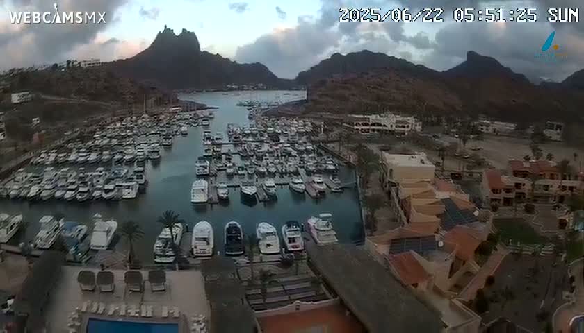A marina filled with numerous boats is nestled between mountains under a partly cloudy sunrise.