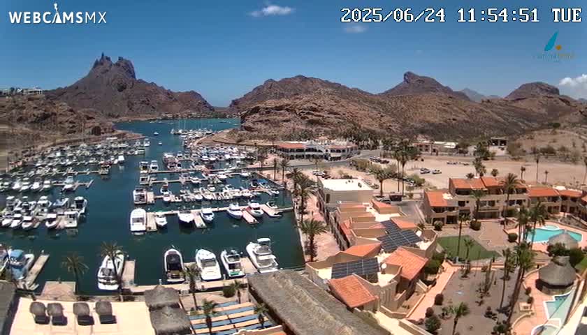 A marina filled with boats is nestled between brown, rocky mountains under a clear, sunny sky.