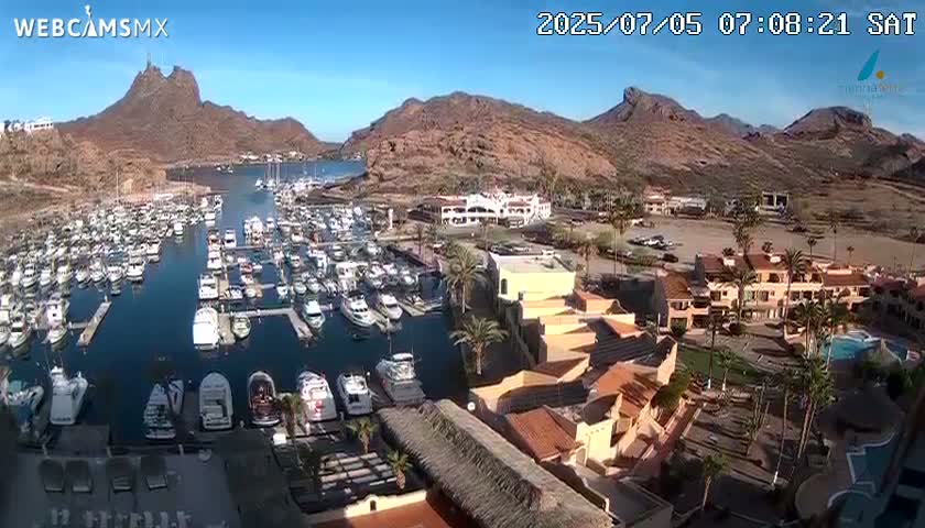 An aerial view shows a marina filled with boats nestled between desert mountains under a clear, sunny sky.
