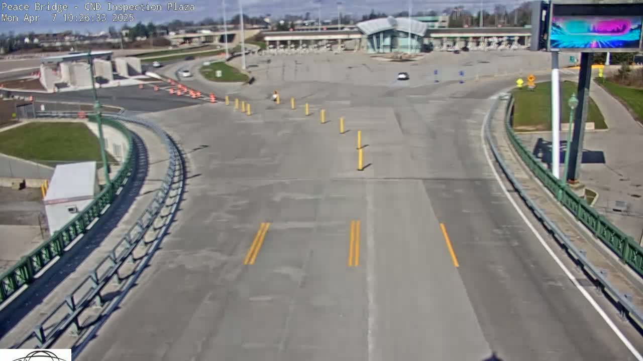 An empty roadway curves toward a border inspection plaza under a sunny sky.