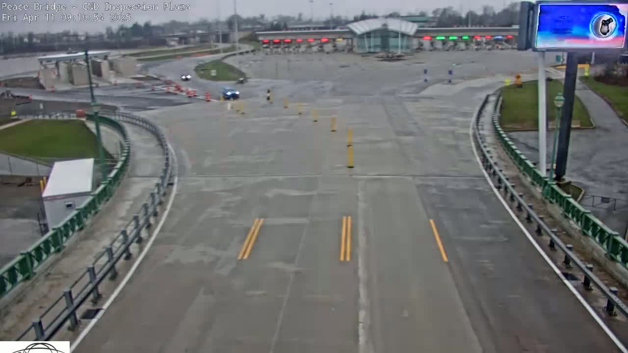 A wet, overcast day at a border crossing inspection plaza shows a mostly empty roadway curving toward a building with several inspection booths.