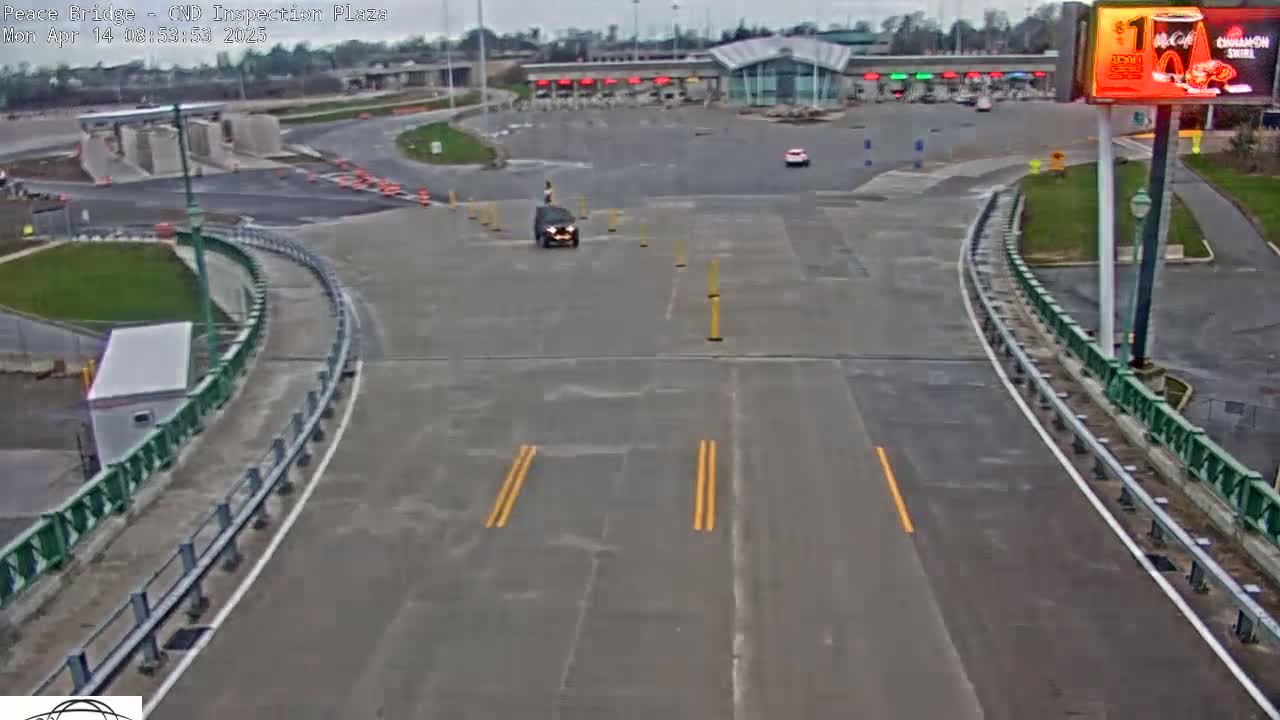 A dark-colored SUV drives toward a mostly empty border inspection plaza on an overcast day.