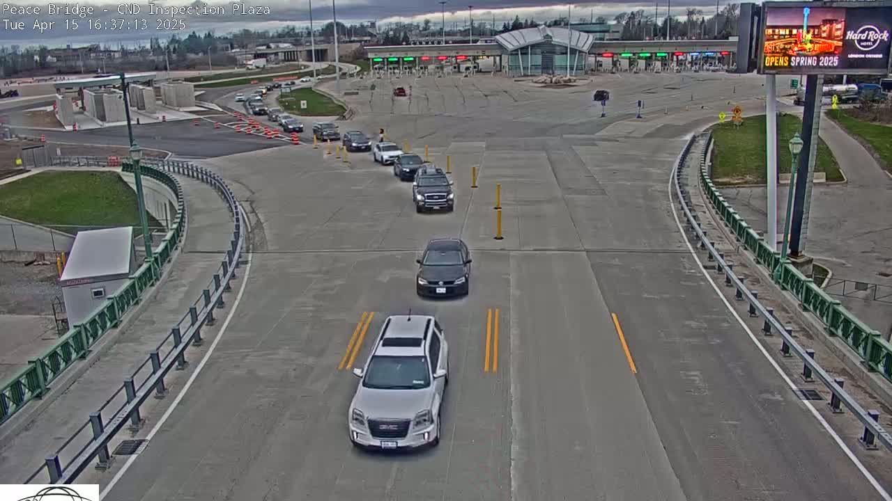 Several vehicles are slowly approaching a border inspection plaza on a mostly cloudy day.