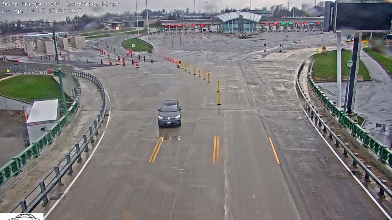 A dark-colored car drives on a wet road curving toward a border inspection plaza under an overcast sky.