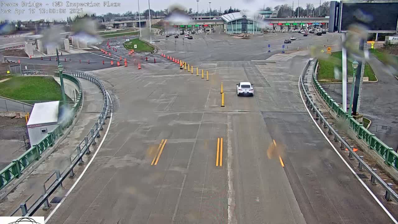 A high-angle, long shot of a roadway curving toward a border inspection plaza on an overcast day, with a single white car driving away from the plaza.