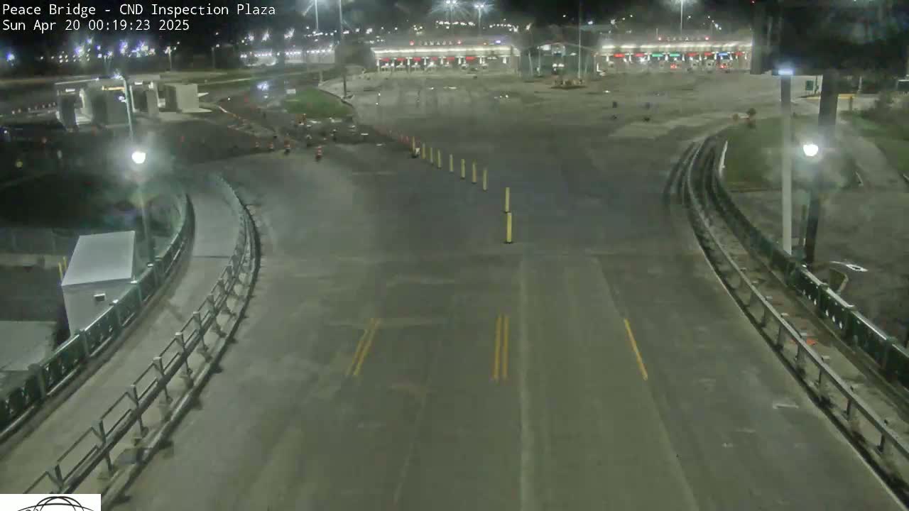 A nighttime overhead view shows an empty roadway curving toward a border crossing inspection plaza under clear skies.