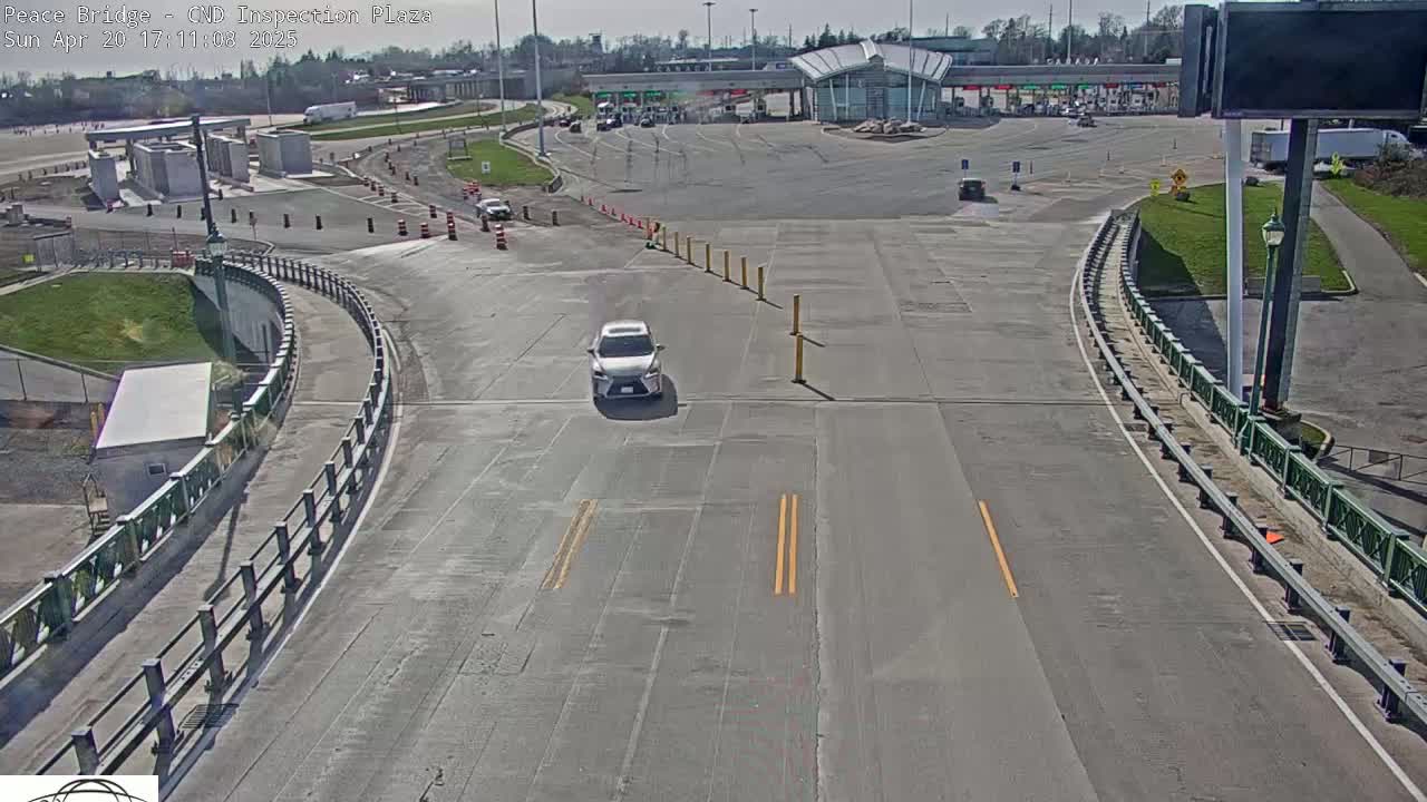 A silver car drives on a roadway curving toward a border inspection plaza on a sunny day.