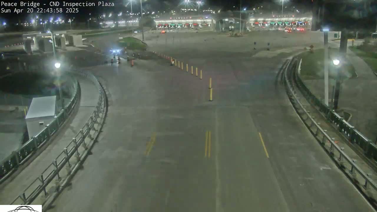 A nighttime, wet road curves toward a border crossing inspection plaza with minimal traffic.