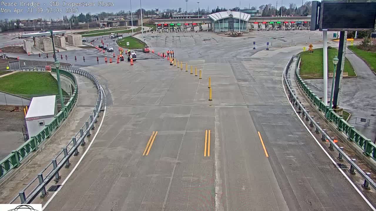 A wet, empty roadway curves toward a border crossing inspection plaza with a few vehicles and workers visible under an overcast sky.