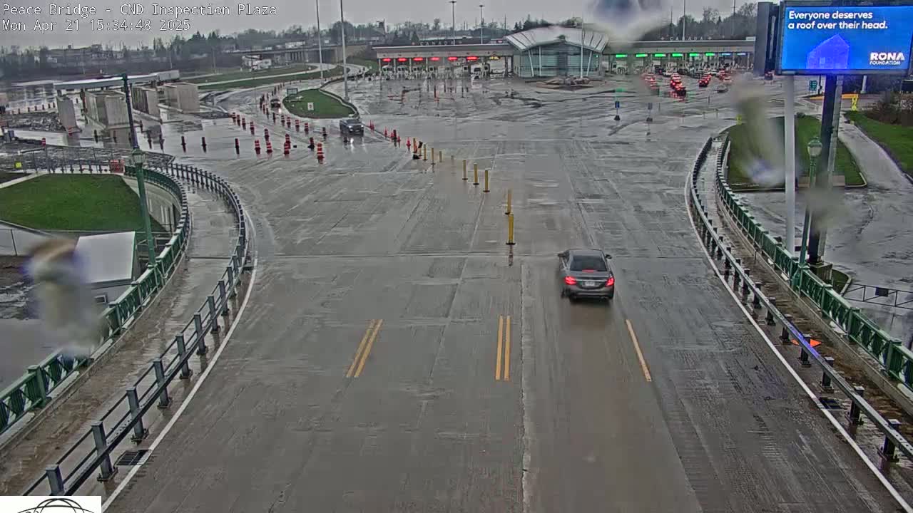 A wet, gray road curves through a border inspection plaza under an overcast sky, with a single car visible on the roadway.