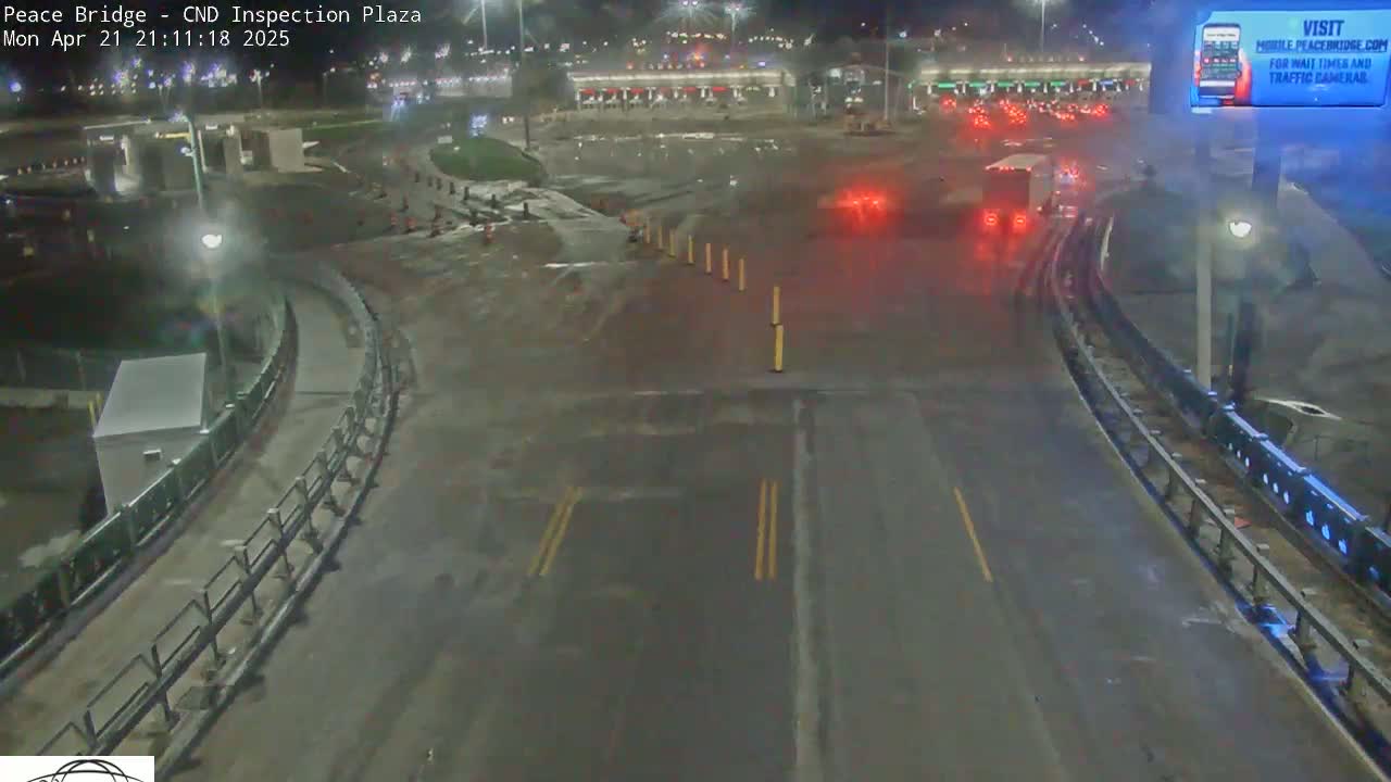 A wet, nighttime view of a mostly empty highway leading to a border inspection plaza with a few vehicles visible in the distance.
