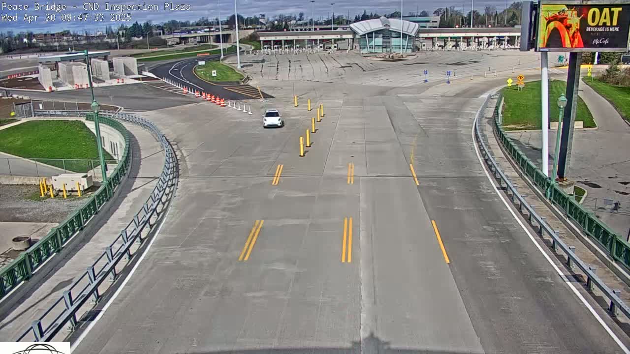 A single white car drives toward a border inspection plaza on a sunny day.