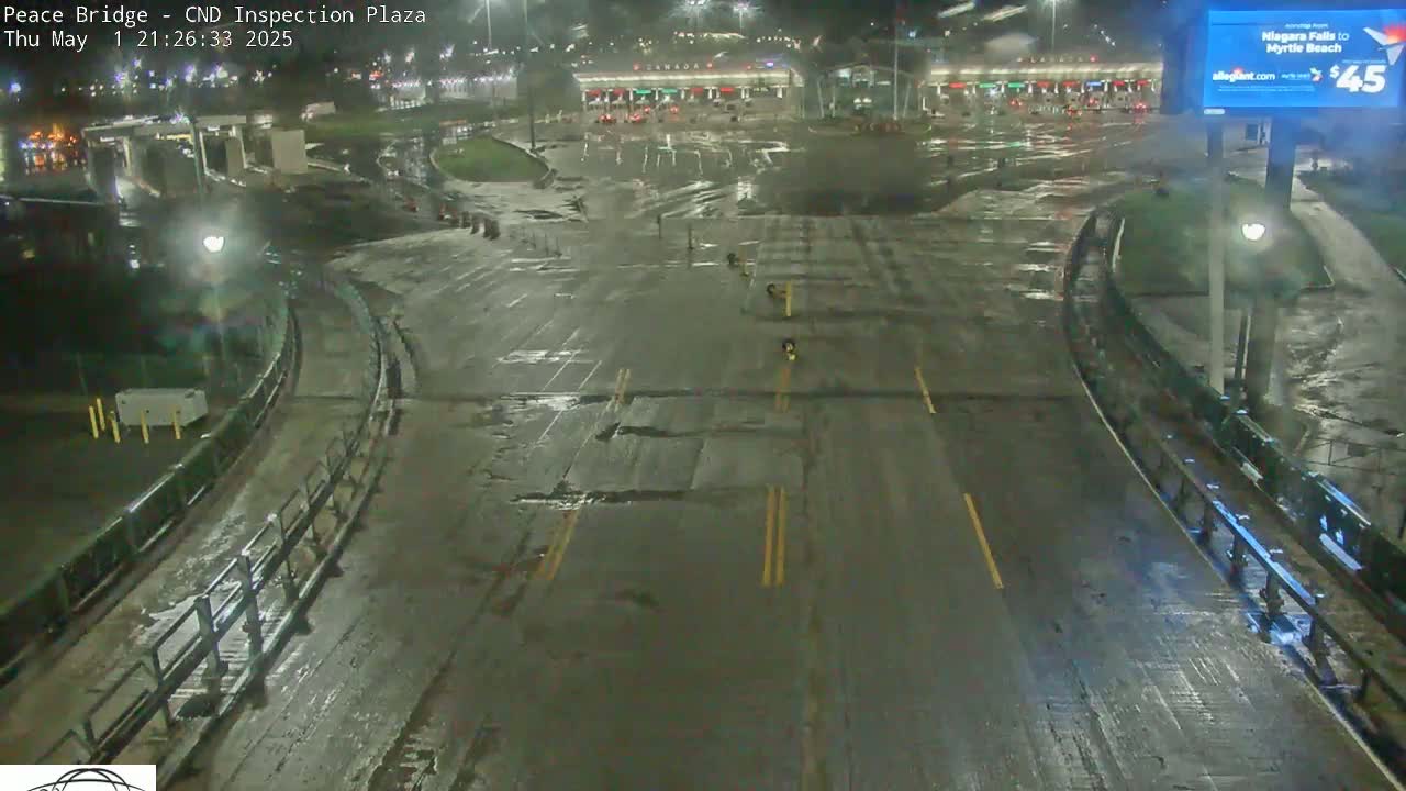 A wet, empty roadway at night curves through a mostly empty border crossing plaza, under dim lighting.