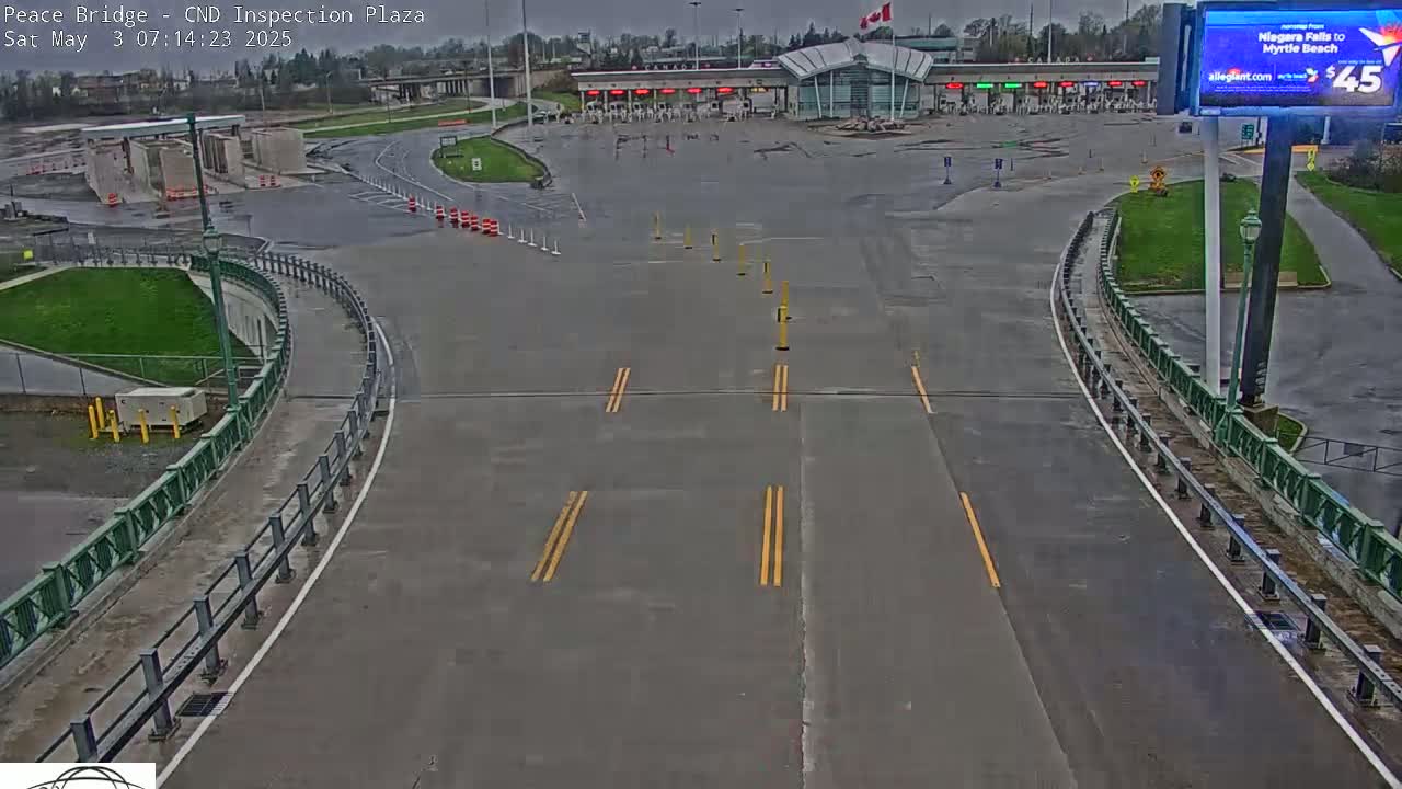 A wet, empty roadway curves toward a border inspection plaza under an overcast sky.