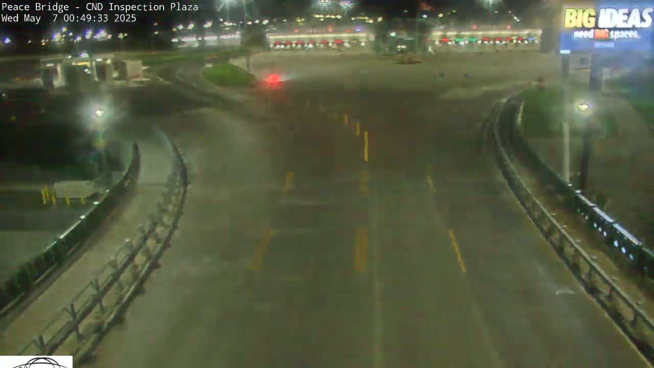 A wet, nighttime view of a roadway curving through a plaza with sparse vegetation and buildings in the distance under dim lighting.