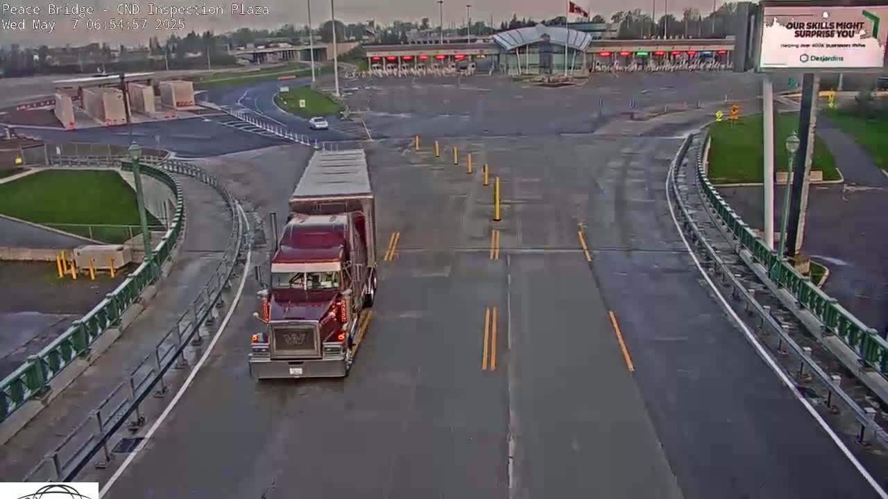 A maroon semi-truck is approaching a border inspection plaza on a wet road under an overcast sky.