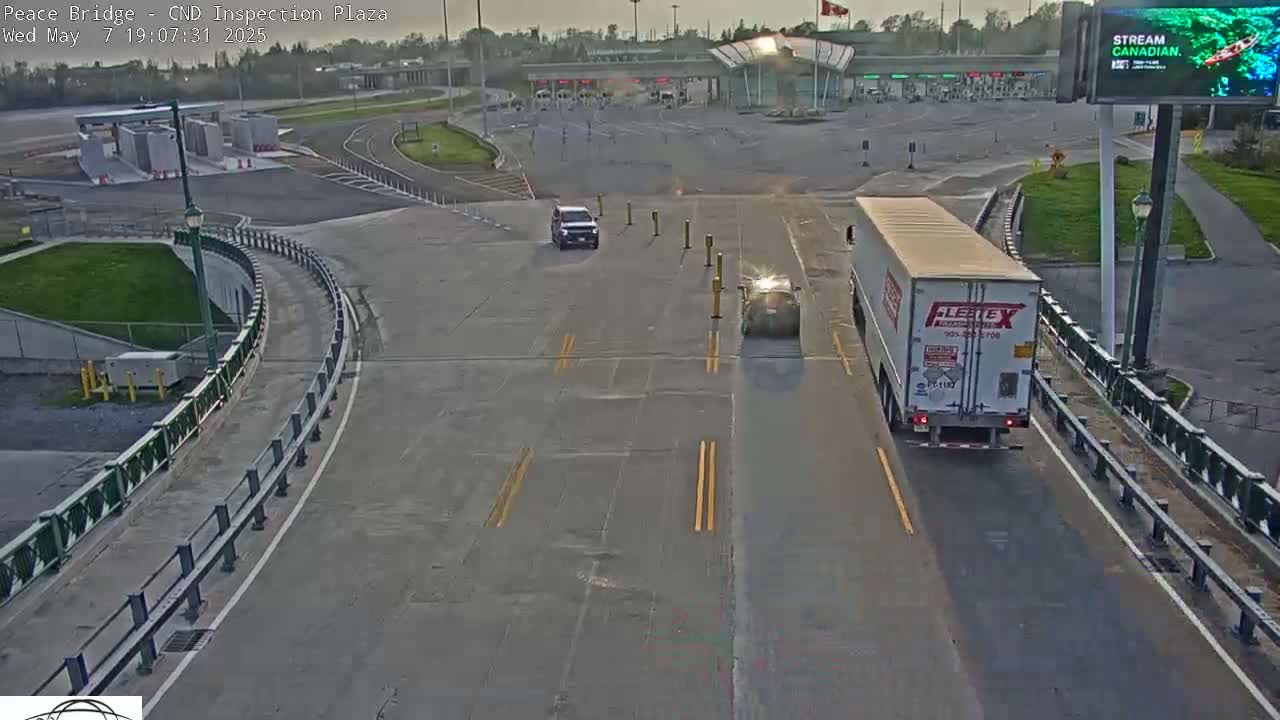 A sunny overhead view of a border crossing inspection plaza shows several vehicles, including a semi-truck, navigating the roadways.