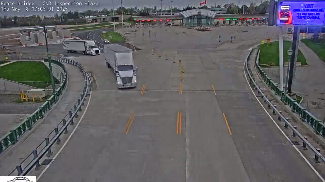 A mostly empty border crossing inspection plaza with two semi-trucks visible on a partly cloudy day.