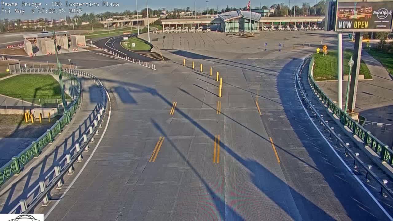 A sunny overhead view of an empty border inspection plaza with curved roadways and long shadows cast by light posts.