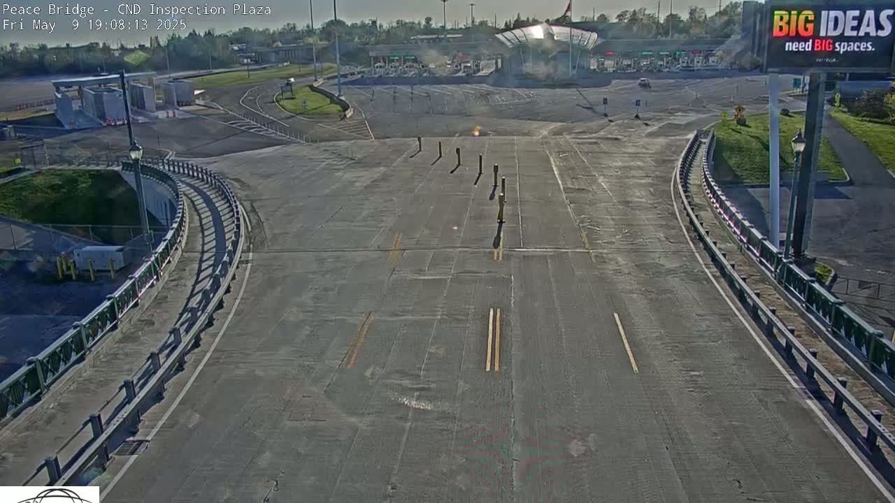 A mostly empty roadway curves toward a border inspection plaza under a sunny sky.