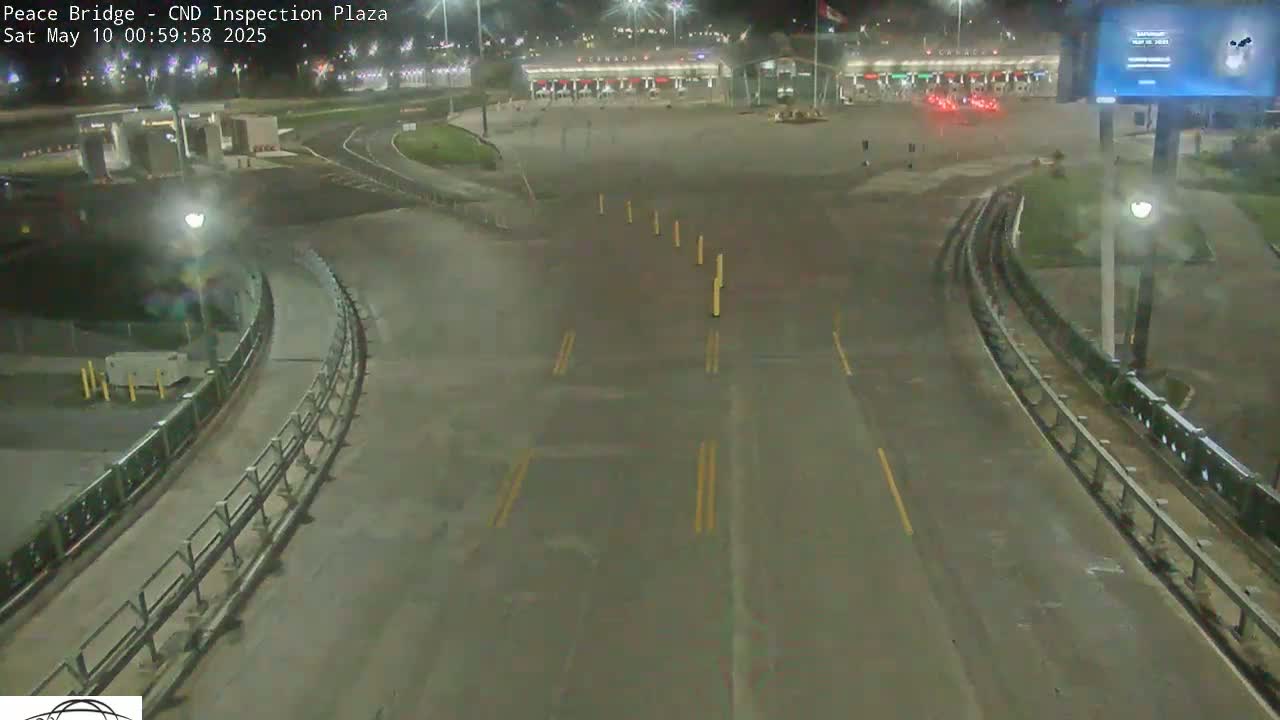 A nighttime overhead view shows a mostly empty border inspection plaza with a roadway curving toward it, under dark skies.