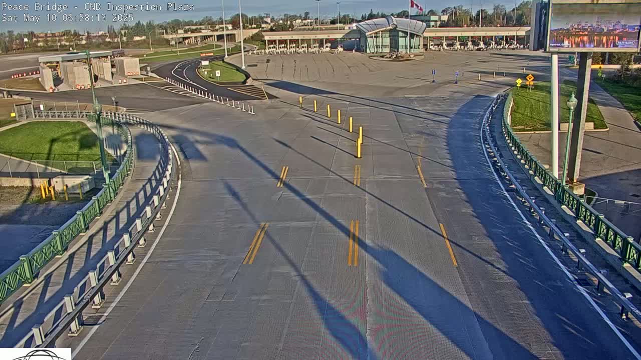 A high-angle view of a mostly empty roadway curving toward a border crossing inspection plaza on a sunny day, with long shadows cast across the pavement.