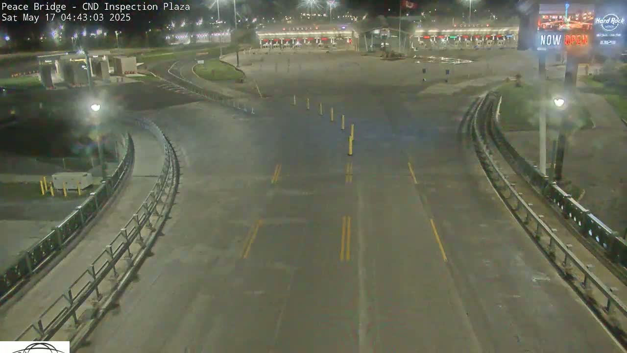 A nighttime, overhead view shows an empty roadway curving toward a border crossing inspection plaza with minimal traffic.