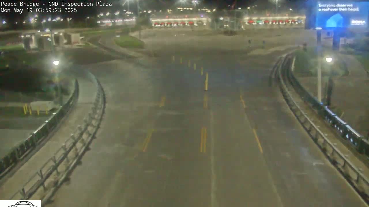 A wet, nighttime view of an empty roadway curving through a plaza with buildings and lights in the background.