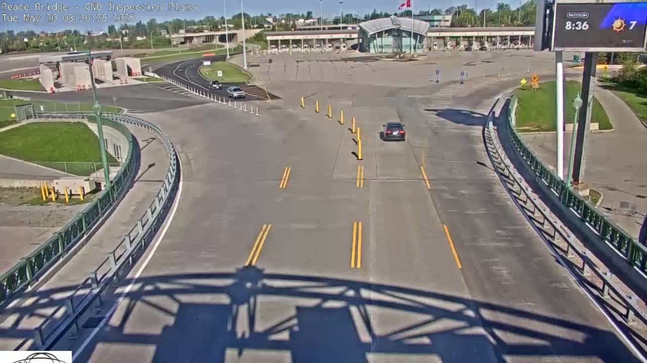 A high-angle, sunny view shows a border inspection plaza with a few vehicles, and a roadway curving toward the viewer.