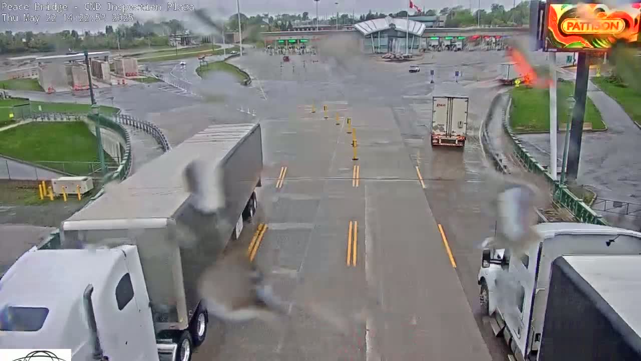 A wet, rainy scene at a border crossing shows several semi-trucks and cars at a customs inspection plaza.
