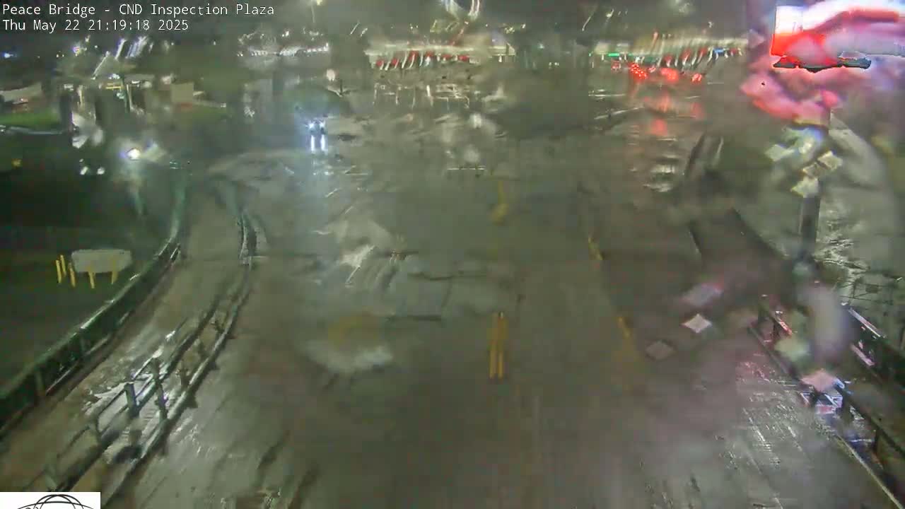 A nighttime view of a rain-soaked roadway and parking lot, with blurry car lights visible in the distance.