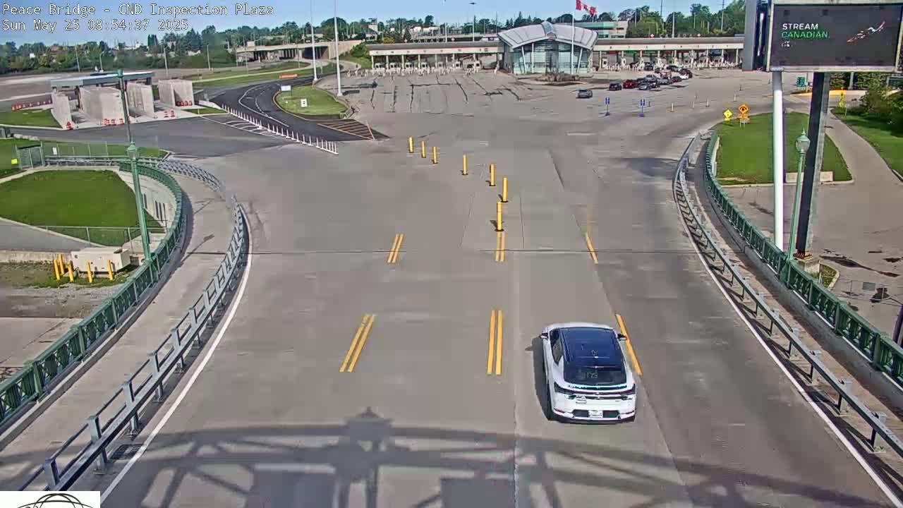 A white car drives toward a mostly empty border inspection plaza on a sunny day.