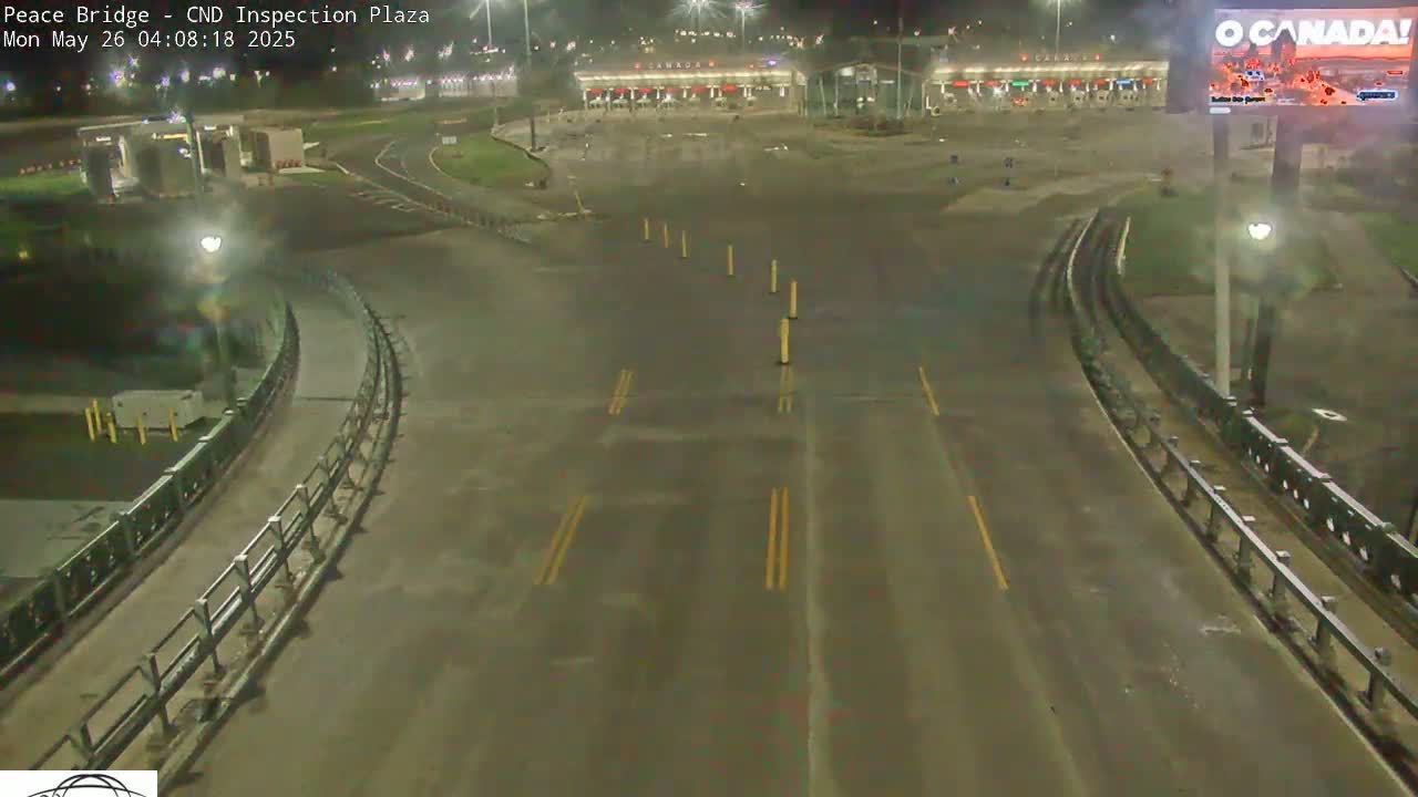 A nighttime, wet roadway curves through an empty border crossing inspection plaza.