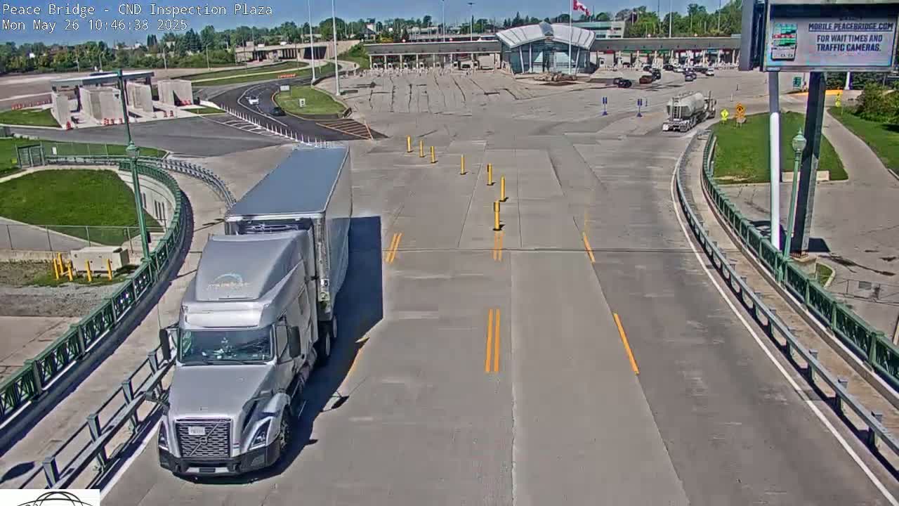 A silver semi-truck approaches a border inspection plaza on a sunny day.