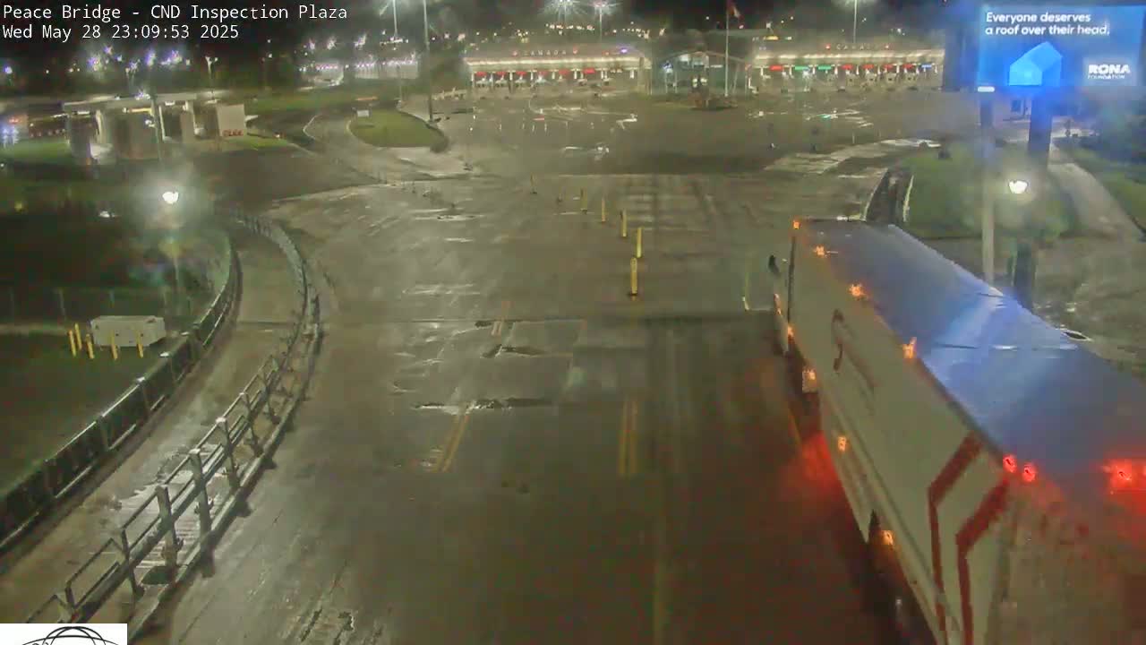 A wet, nighttime view of a border inspection plaza with a large truck in the foreground and a mostly empty plaza in the background.
