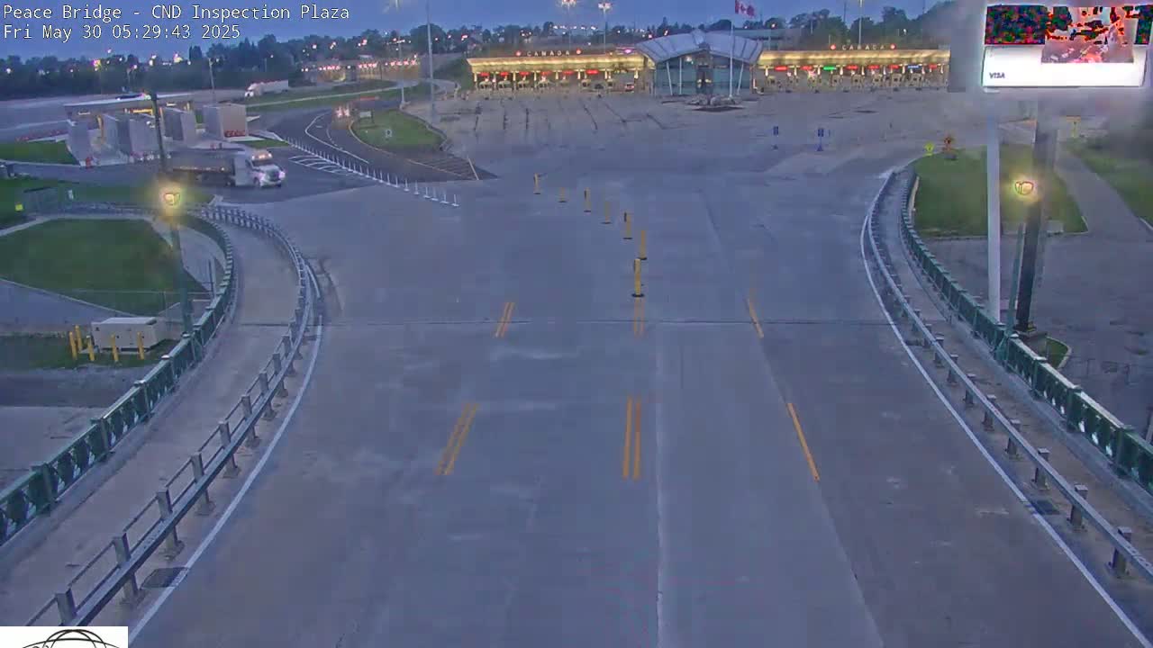 A semi-truck drives toward a mostly empty Canadian border inspection plaza under a twilight sky.