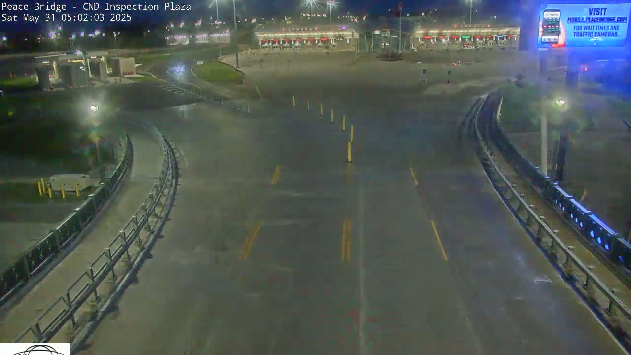 A nighttime, wet road curves toward a border crossing inspection plaza with minimal traffic.